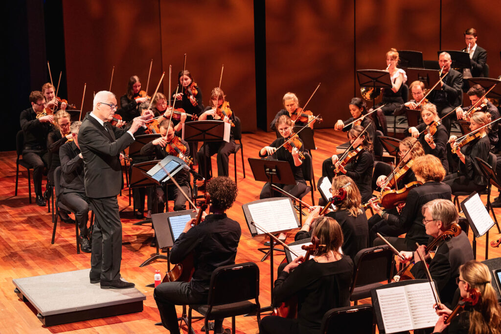 Strijkers van het Flevolands Philharmonisch Orkest maken muziek op een podium met dirigent Hans Welle