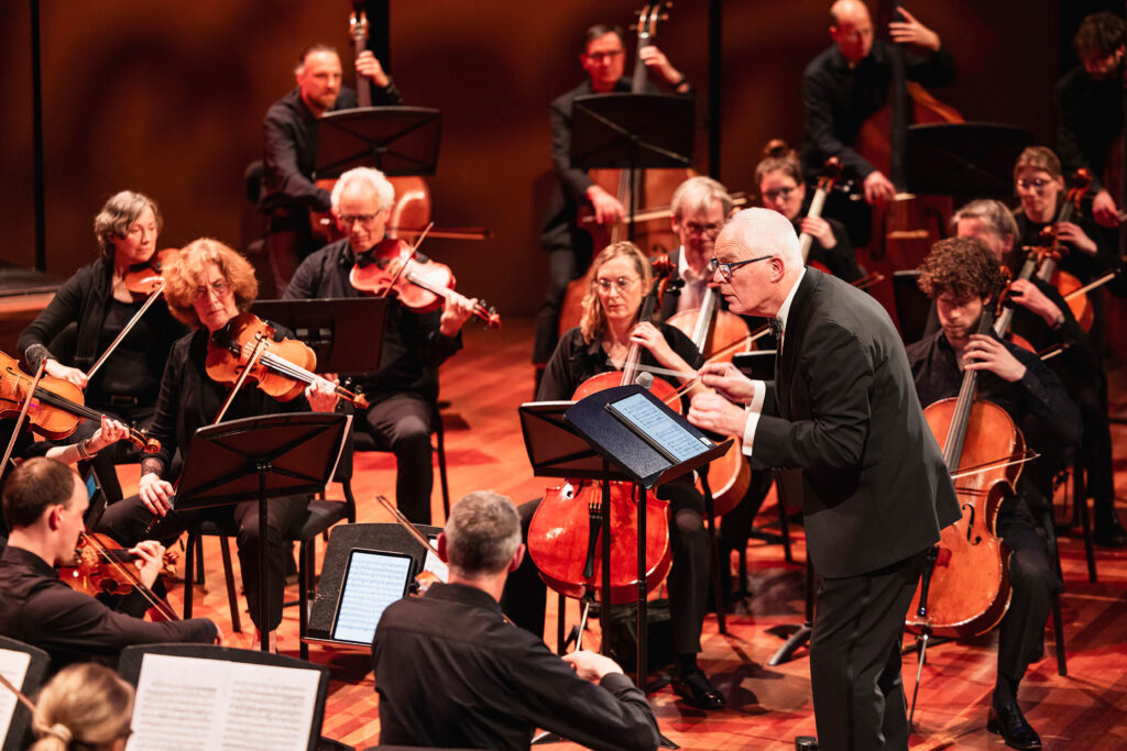 Strijkers van het Flevolands Philharmonisch Orkest maken muziek op een podium met dirigent Hans Welle